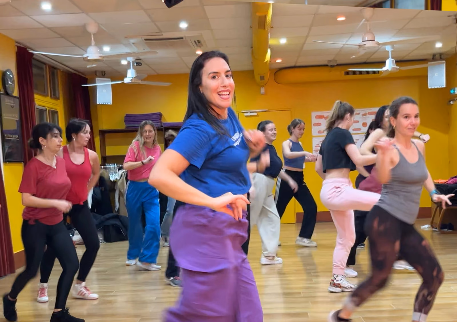 Une professeure en tee-shirt bleu mène un groupe qui danse face au miroir, salle jaune.