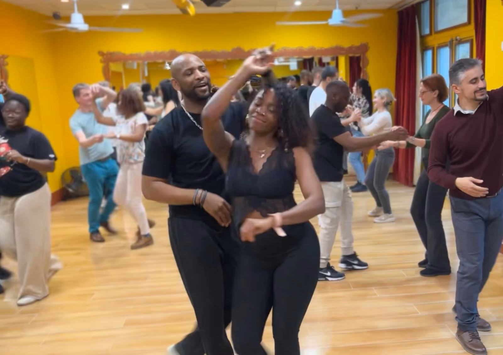 Pendant une journée portes ouvertes, un couple danse au premier plan d’une salle jaune pleine.