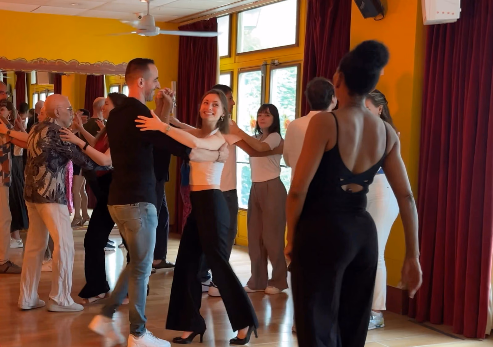 Des couples de tous âges dansent pendant un cours en salle jaune, rideaux rouges et miroirs au fond.